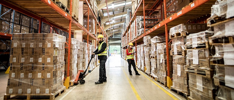 Man and woman wearing hardhats and high-vis jackets stood in a warehouse surrounded by pallets of boxes