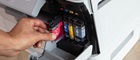 A hand placing a magenta ink cartridge into a Brother printer next to cyan, yellow, and black cartridges