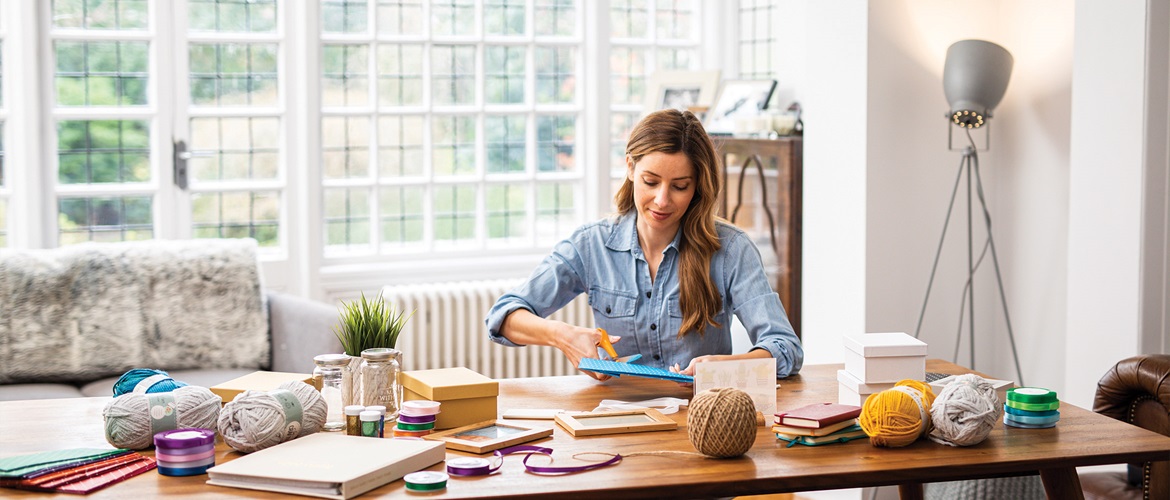 Woman using scissors sat at a table covered with boxes, jars, balls of wool and folders