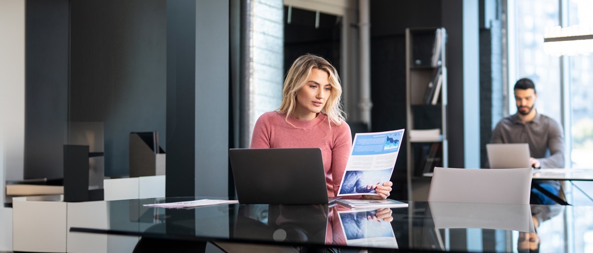 A man and woman working together in a modern office, with one reviewing a printed document and the other working on a laptop