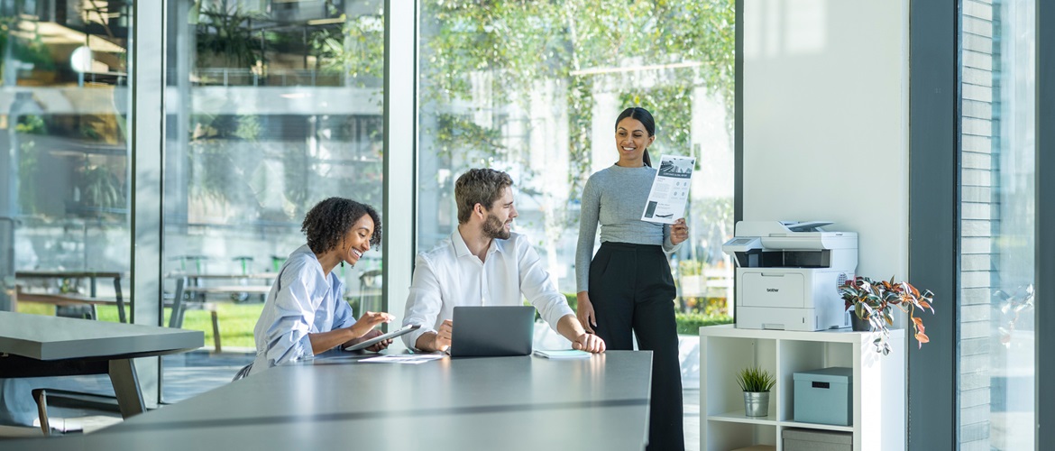 Three people in an office, one standing with a document next to a printer and two seated with laptops.