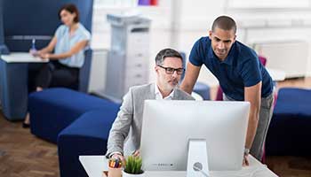 Man with glasses sat at desk, man stood looking over shoulders at monitor, woman sat at a booth, printer, plant, pens